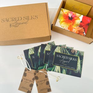 May include: A brown cardboard box and an open box with a floral card and a handwritten note. Several business cards with a palm leaf design and two hang tags are also visible. The text on the box reads "SACRED SILKS Apparel."