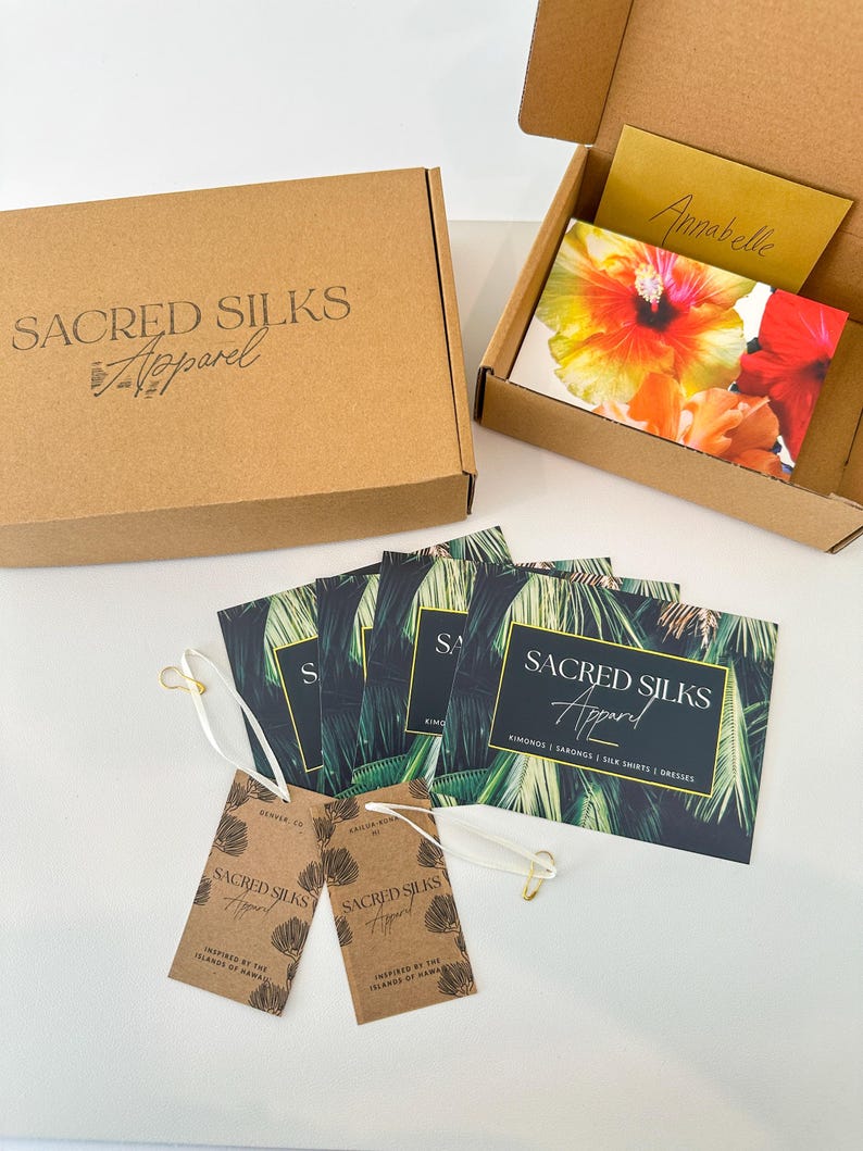 May include: A brown cardboard box and an open box with a floral card and a handwritten note. Several business cards with a palm leaf design and two hang tags are also visible. The text on the box reads "SACRED SILKS Apparel."