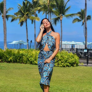 May include: A blue and black paisley print halter top and midi skirt set. The outfit has a keyhole cutout and is worn with beige sandals. The background features palm trees, green grass, and a blue sky.
