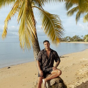 Puede incluir: Una persona con una camisa de manga corta estampada oscura y pantalones cortos negros est&aacute; sentada en el tronco de una palmera en una playa de arena. El fondo muestra el mar, el cielo y palmeras.