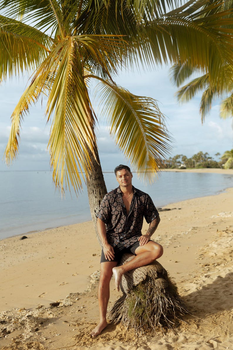 Puede incluir: Una persona con una camisa de manga corta estampada oscura y pantalones cortos negros est&aacute; sentada en el tronco de una palmera en una playa de arena. El fondo muestra el mar, el cielo y palmeras.
