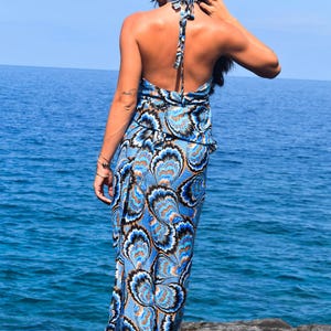 May include: A halter-neck maxi dress with a blue, black, and orange floral pattern. The dress has an open back and is worn near the ocean. The dress is a summer or beachwear item.