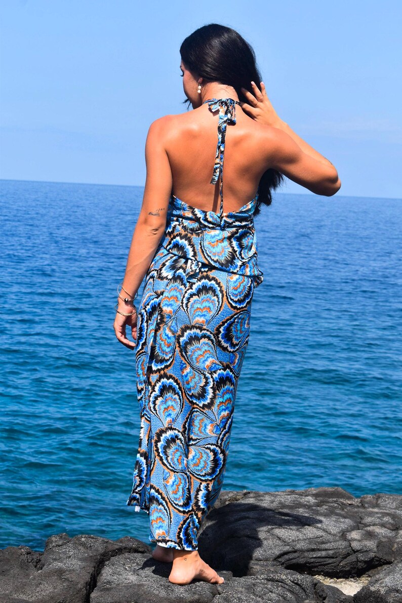 May include: A halter-neck maxi dress with a blue, black, and orange floral pattern. The dress has an open back and is worn near the ocean. The dress is a summer or beachwear item.