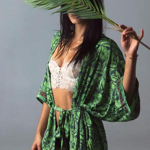 Puede incluir: Bata estilo kimono con estampado floral verde y azul con corbata a juego. La bata se lleva sobre un bralette de encaje blanco. Una hoja de palma se sostiene frente a la cara.