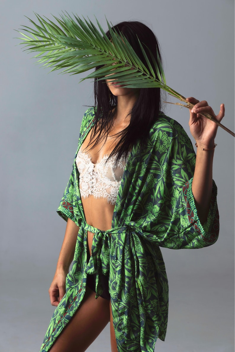 Puede incluir: Bata estilo kimono con estampado floral verde y azul con corbata a juego. La bata se lleva sobre un bralette de encaje blanco. Una hoja de palma se sostiene frente a la cara.