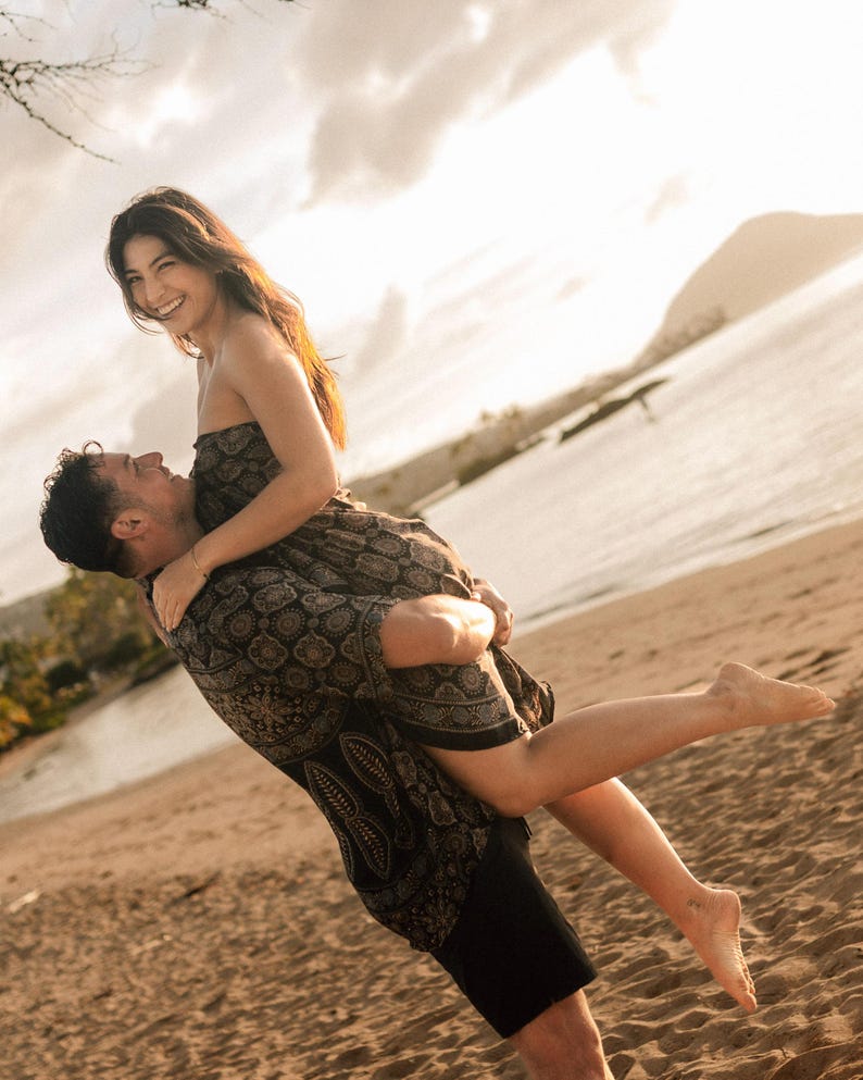 Puede incluir: Una persona lleva a una mujer en una playa de arena. La mujer lleva un vestido oscuro sin tirantes con un estampado floral repetido. El fondo muestra el mar y un cielo nublado.