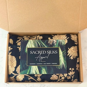 Pu&ograve; includere: Una scatola di spedizione in cartone contenente un tessuto nero piegato con un motivo floreale dorato. Sopra &egrave; appoggiato un cartoncino rettangolare con sfondo di foglie di palma, il testo SACRED SILKS Apparel e le parole KIMONOS | SARONGS | SILK SHIRTS | DRESSES.