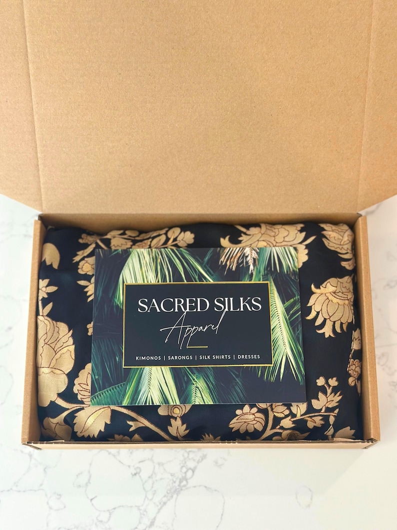 Pu&ograve; includere: Una scatola di spedizione in cartone contenente un tessuto nero piegato con un motivo floreale dorato. Sopra &egrave; appoggiato un cartoncino rettangolare con sfondo di foglie di palma, il testo SACRED SILKS Apparel e le parole KIMONOS | SARONGS | SILK SHIRTS | DRESSES.