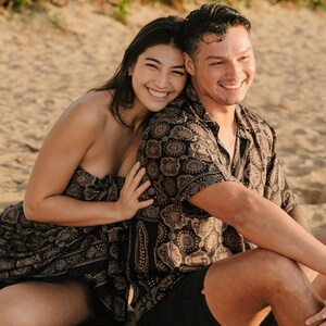Puede incluir: Una pareja sonriente est&aacute; sentada en una playa arenosa. La mujer lleva un vestido negro sin tirantes con estampado. El hombre lleva una camisa de manga corta con botones con un patr&oacute;n similar y pantalones cortos negros. El fondo muestra arena y vegetaci&oacute;n.
