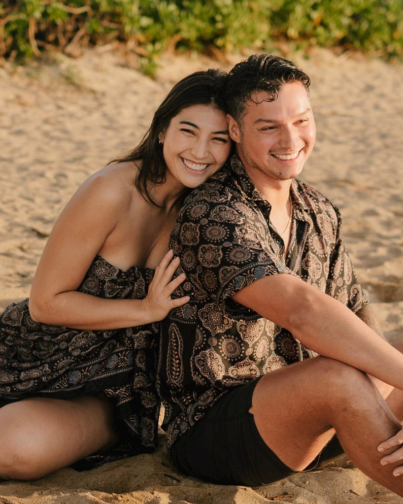 Puede incluir: Una pareja sonriente est&aacute; sentada en una playa arenosa. La mujer lleva un vestido negro sin tirantes con estampado. El hombre lleva una camisa de manga corta con botones con un patr&oacute;n similar y pantalones cortos negros. El fondo muestra arena y vegetaci&oacute;n.