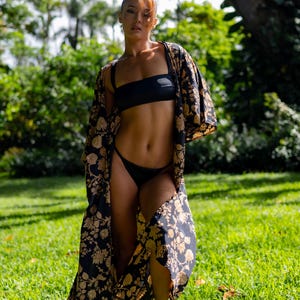 Pu&ograve; includere: Un set bikini nero composto da un top a fascia e uno slip a taglio alto, abbinato a un lungo kimono aperto sul davanti con un motivo floreale dorato ripetuto su sfondo nero, maniche ampie e una silhouette fluida che arriva fino a terra.