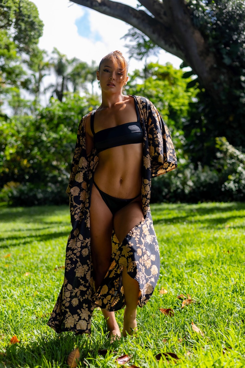 Pu&ograve; includere: Un set bikini nero composto da un top a fascia e uno slip a taglio alto, abbinato a un lungo kimono aperto sul davanti con un motivo floreale dorato ripetuto su sfondo nero, maniche ampie e una silhouette fluida che arriva fino a terra.