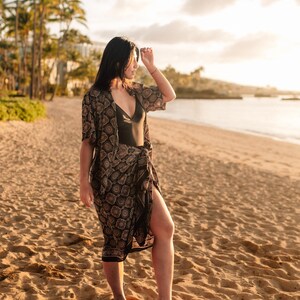 Puede incluir: Una persona en una playa de arena con un traje de ba&ntilde;o negro, un kimono estampado y una falda envolvente a juego. El kimono y la falda tienen un dise&ntilde;o floral repetido en negro y marr&oacute;n. La escena est&aacute; iluminada por la c&aacute;lida luz del sol.