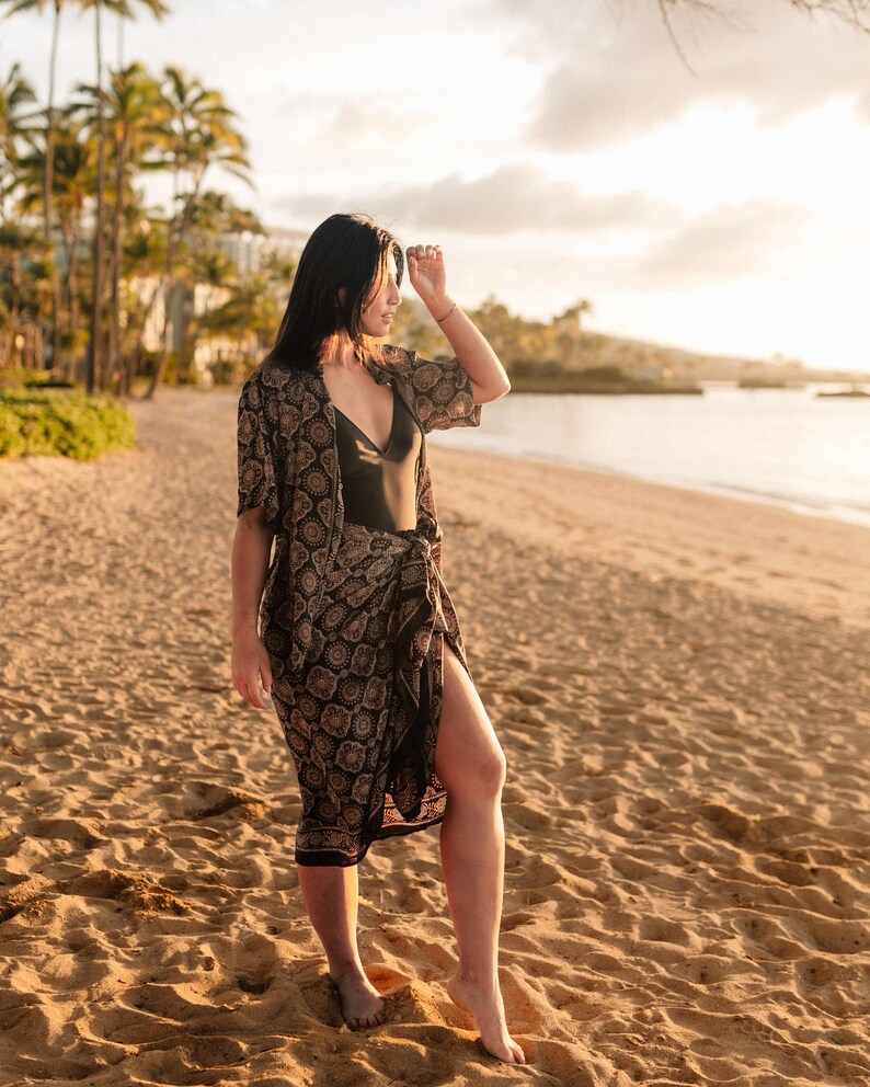 Puede incluir: Una persona en una playa de arena con un traje de ba&ntilde;o negro, un kimono estampado y una falda envolvente a juego. El kimono y la falda tienen un dise&ntilde;o floral repetido en negro y marr&oacute;n. La escena est&aacute; iluminada por la c&aacute;lida luz del sol.
