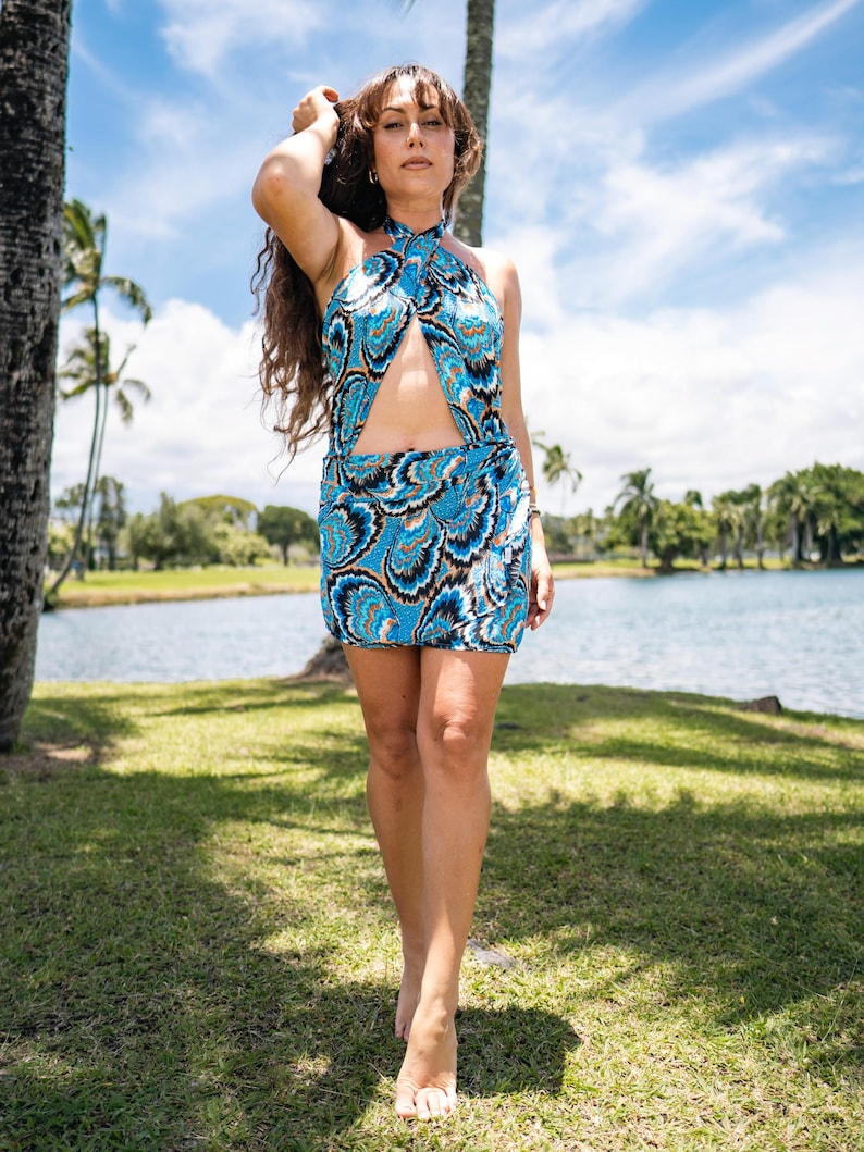 May include: A halter-neck mini dress with a blue, black, and white paisley pattern. The dress features a keyhole cutout at the chest and a matching skirt. The model is standing barefoot on grass near a lake, with palm trees and a blue sky in the background.