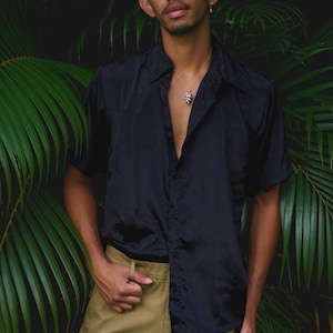 May include: A short-sleeved black button-down shirt with a subtle sheen, paired with tan trousers. A thin gold chain necklace features a small, black and white patterned pendant that hangs at the centre of the chest, resting against the skin.