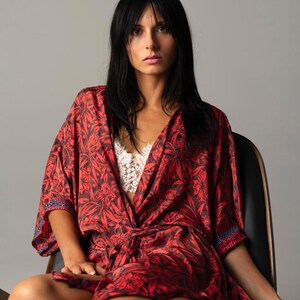 May include: A red and black floral print kimono robe with wide sleeves and a matching tie belt, worn over a white lace bralette. The robe features a dark botanical pattern throughout and a thin blue and white geometric trim along the sleeve edges.
