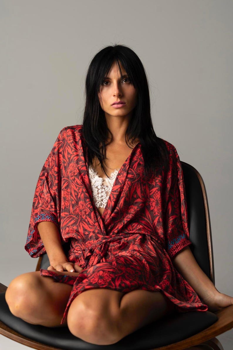 May include: A red and black floral print kimono robe with wide sleeves and a matching tie belt, worn over a white lace bralette. The robe features a dark botanical pattern throughout and a thin blue and white geometric trim along the sleeve edges.