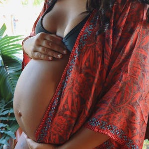 May include: A lightweight, open-front kimono robe featuring a vibrant red and dark brown botanical leaf pattern with a contrasting blue and red geometric border along the edges. The garment is worn over a solid black bikini top, showcasing a loose fit.