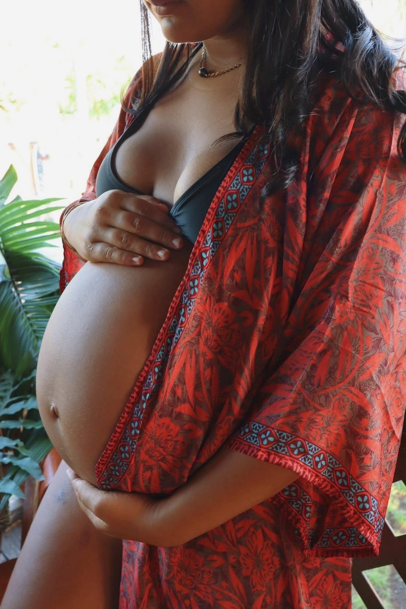 May include: A lightweight, open-front kimono robe featuring a vibrant red and dark brown botanical leaf pattern with a contrasting blue and red geometric border along the edges. The garment is worn over a solid black bikini top, showcasing a loose fit.