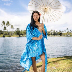 May include: A bright blue kimono-style robe featuring a white and blue marbled pattern with wide sleeves and an open front, paired with a large white paper parasol with visible bamboo ribs, held overhead against a bright outdoor sky.