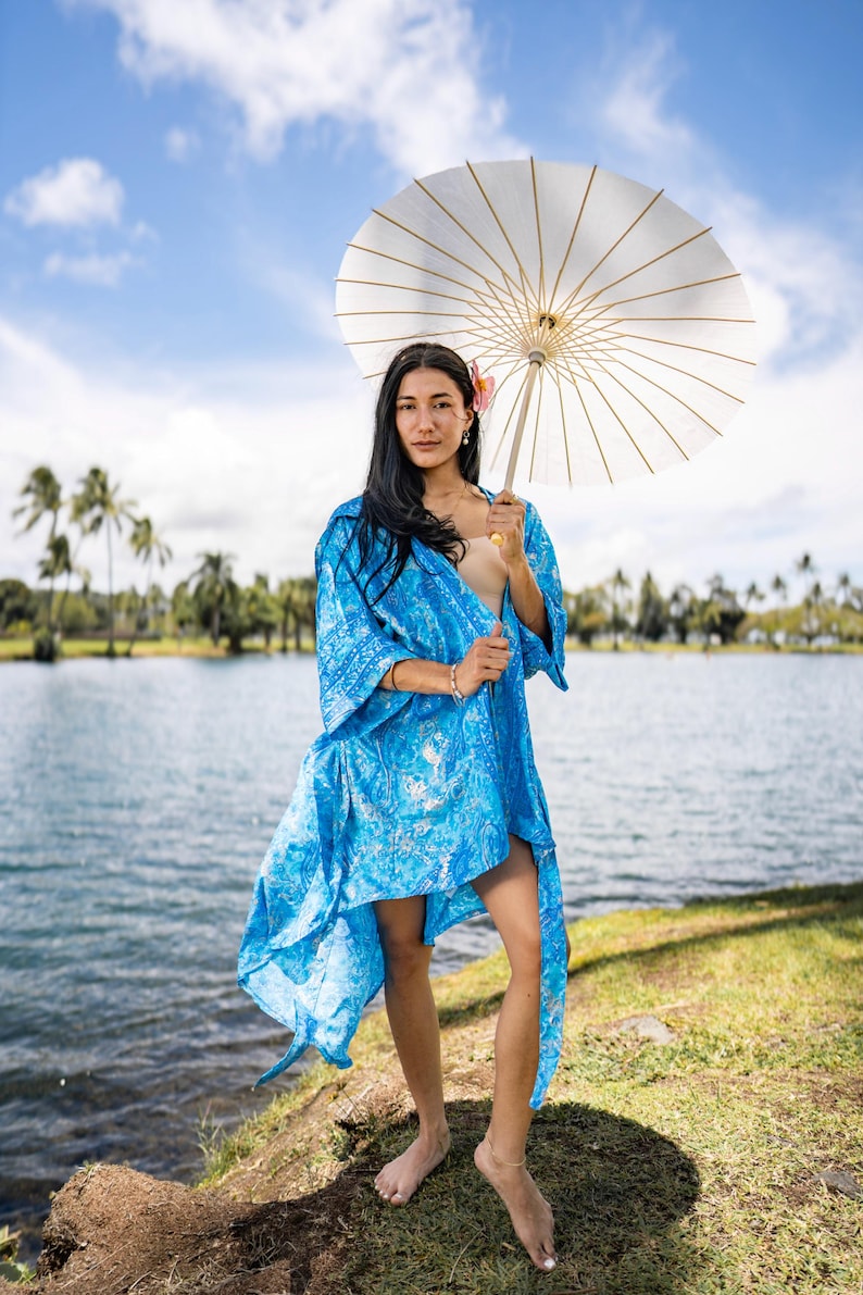 May include: A bright blue kimono-style robe featuring a white and blue marbled pattern with wide sleeves and an open front, paired with a large white paper parasol with visible bamboo ribs, held overhead against a bright outdoor sky.