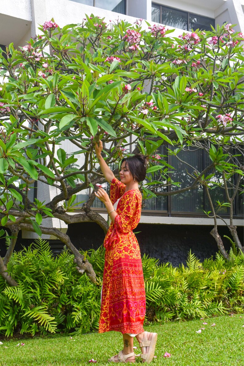 May include: A woman in a long, flowing red and yellow patterned dress stands near a tree with pink flowers. The dress has a vibrant floral design. The scene is set outdoors with green foliage and a building in the background.