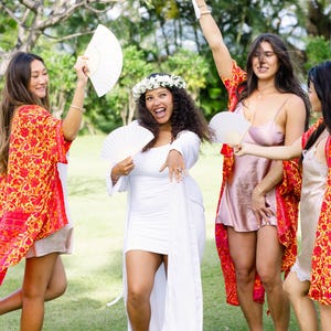 May include: A group of people celebrating outdoors, wearing a variety of outfits. One person is in a white dress with a floral crown, while others wear robes with a red and yellow floral pattern. They are holding white fans.