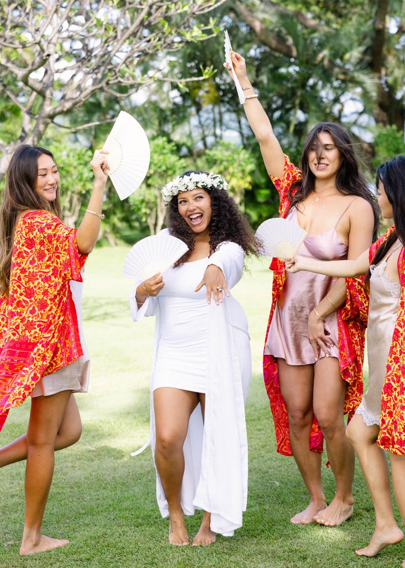 May include: A group of people celebrating outdoors, wearing a variety of outfits. One person is in a white dress with a floral crown, while others wear robes with a red and yellow floral pattern. They are holding white fans.