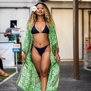 Puede incluir: Una mujer con un bikini negro y un kimono verde con estampado floral. Tiene una flor blanca en el pelo. El kimono tiene un fondo verde con un estampado de hojas y ribetes naranjas y marrones. Camina sobre una superficie gris.