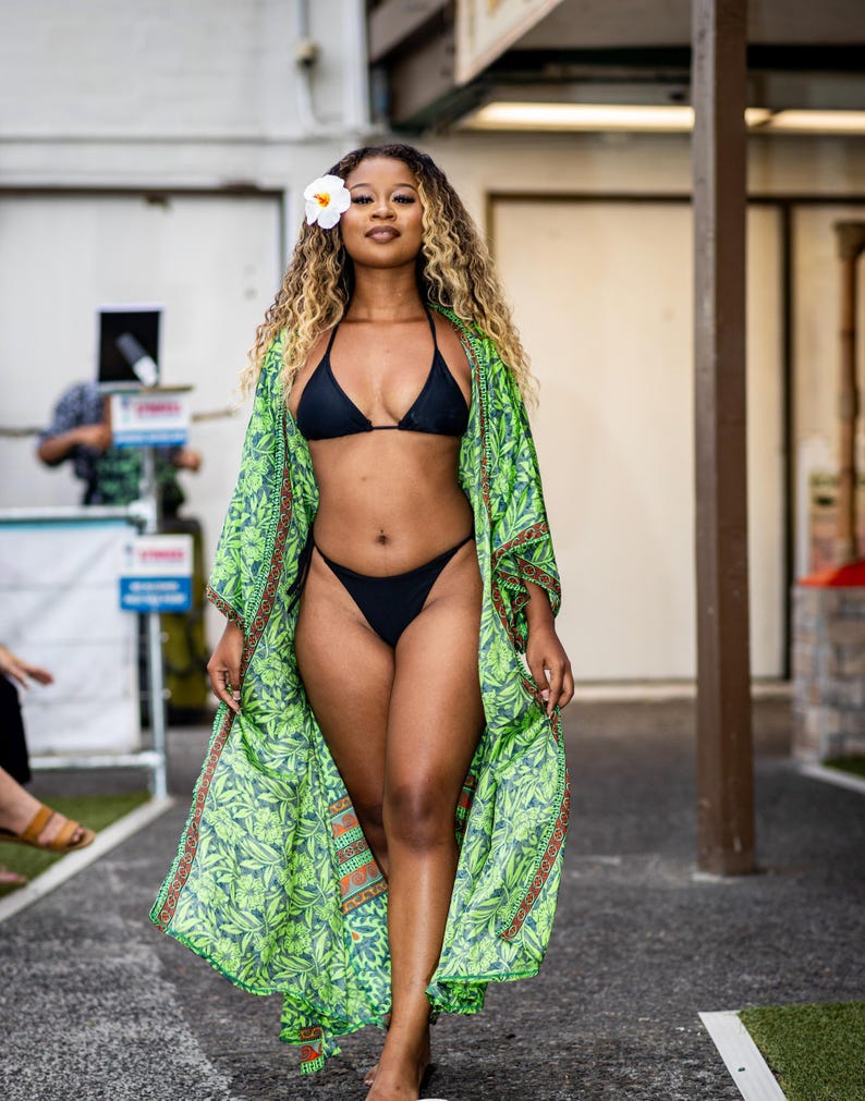 Puede incluir: Una mujer con un bikini negro y un kimono verde con estampado floral. Tiene una flor blanca en el pelo. El kimono tiene un fondo verde con un estampado de hojas y ribetes naranjas y marrones. Camina sobre una superficie gris.