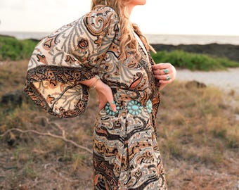 May include: A long-sleeved kimono-style robe featuring a detailed paisley pattern in shades of black, tan, and orange. The garment is cinched at the waist with a belt composed of multiple oval-shaped turquoise stones set in silver metal.