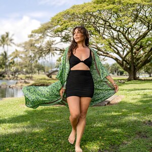 May include: A black two-piece outfit consisting of a knotted crop top and a fitted mini skirt, paired with a long, open-front kimono featuring a vibrant green botanical leaf print and a decorative patterned border along the edges.