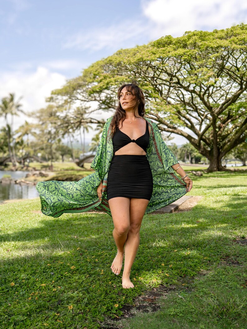 May include: A black two-piece outfit consisting of a knotted crop top and a fitted mini skirt, paired with a long, open-front kimono featuring a vibrant green botanical leaf print and a decorative patterned border along the edges.