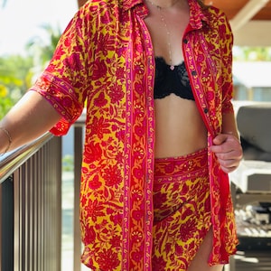 May include: A button-down shirt and matching wrap skirt set featuring a vibrant floral pattern in shades of bright yellow, hot pink, and deep red. The shirt has short sleeves and a collar, while the skirt features a side slit design.