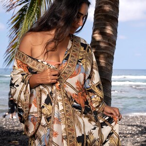 May include: A long, flowing kimono-style robe featuring a cream-colored base with a repeating pattern of palm trees and tropical foliage in shades of orange, black, and blue. The garment has wide sleeves and a decorative border with geometric motifs.