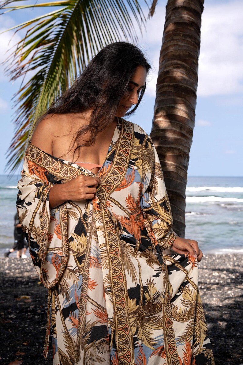 May include: A long, flowing kimono-style robe featuring a cream-colored base with a repeating pattern of palm trees and tropical foliage in shades of orange, black, and blue. The garment has wide sleeves and a decorative border with geometric motifs.