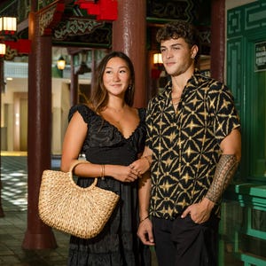 Puede incluir: Un vestido negro con volantes y cuerpo fruncido junto a un bolso de mano de paja trenzada natural con asas redondas, y una camisa negra de manga corta con botones y estampado de estrellas geométricas doradas con pantalones cortos negros.