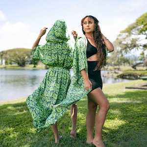 Puede incluir: Dos personas de pie sobre hierba cerca de un lago. Una persona lleva un vestido con capucha con estampado floral verde y azul marino. La otra persona lleva un top negro, una falda negra y una sobrecamisa tipo kimono con estampado floral verde y azul marino.