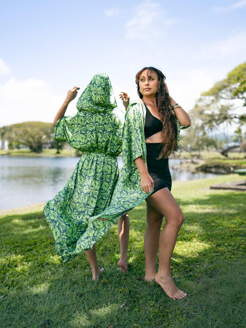 Puede incluir: Dos personas de pie sobre hierba cerca de un lago. Una persona lleva un vestido con capucha con estampado floral verde y azul marino. La otra persona lleva un top negro, una falda negra y una sobrecamisa tipo kimono con estampado floral verde y azul marino.