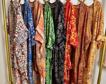 May include: A collection of eight long, open-front robes hanging on wooden hangers on a gold metal rack. The robes feature various colourful patterns, including floral, paisley, and geometric designs in shades of cream, blue, orange, green, black, red, and yellow.
