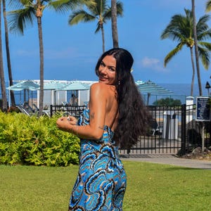 May include: A blue and white patterned jumpsuit with a halter neckline. The jumpsuit features a swirling design in shades of blue, white, and brown. The background includes palm trees, green grass, and a blue sky.