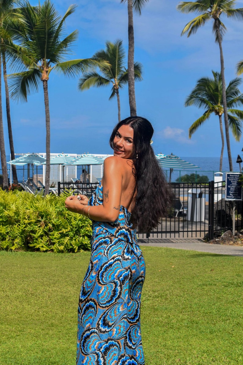 May include: A blue and white patterned jumpsuit with a halter neckline. The jumpsuit features a swirling design in shades of blue, white, and brown. The background includes palm trees, green grass, and a blue sky.