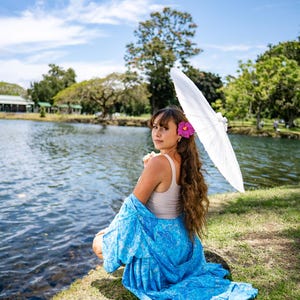 May include: A beige tank top paired with a vibrant blue patterned wrap or sarong featuring intricate white designs. The ensemble is accessorized with a bright pink hibiscus flower tucked into long wavy hair and a white paper parasol held overhead.