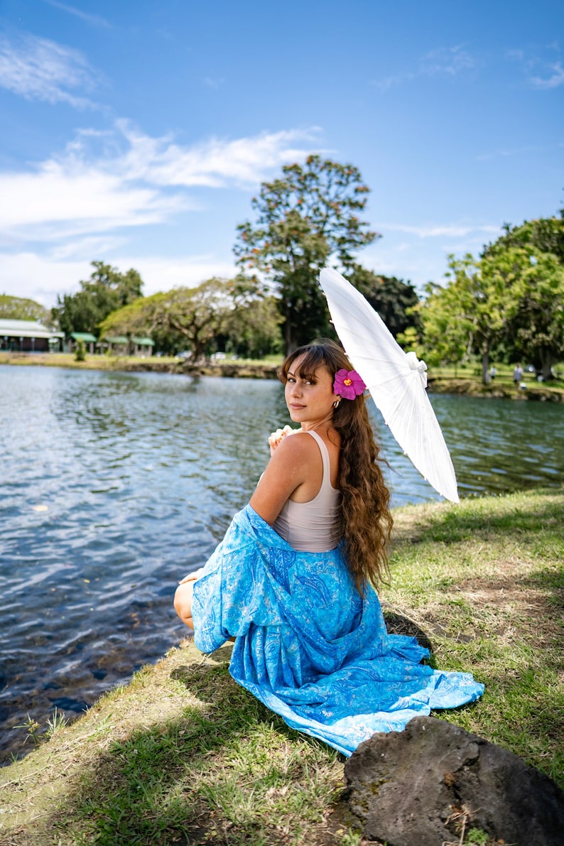 May include: A beige tank top paired with a vibrant blue patterned wrap or sarong featuring intricate white designs. The ensemble is accessorized with a bright pink hibiscus flower tucked into long wavy hair and a white paper parasol held overhead.