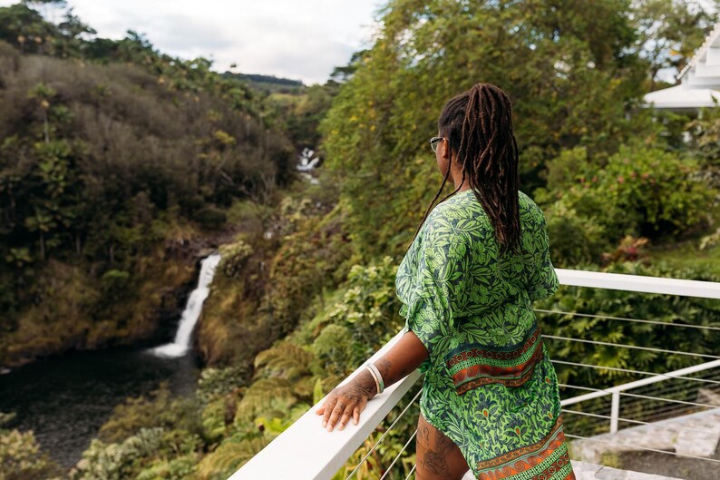 Puede incluir: Una persona con una parte superior estampada verde con acentos naranjas y marrones, de pie en un balc&oacute;n blanco con vista a una cascada y un exuberante paisaje verde. La persona lleva gafas y tiene largas rastas.