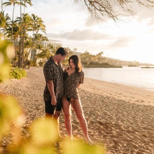 Puede incluir: Una pareja en una playa de arena, ambos con camisas de manga corta a juego con un patr&oacute;n geom&eacute;trico. El hombre lleva pantalones cortos negros y la mujer un traje de ba&ntilde;o negro. Se ven palmeras y un edificio al fondo.