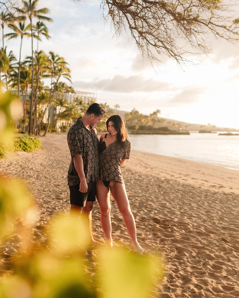 Puede incluir: Una pareja en una playa de arena, ambos con camisas de manga corta a juego con un patr&oacute;n geom&eacute;trico. El hombre lleva pantalones cortos negros y la mujer un traje de ba&ntilde;o negro. Se ven palmeras y un edificio al fondo.