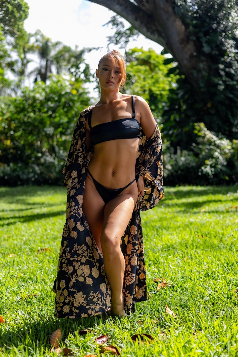 Pu&ograve; includere: Un set bikini nero composto da un top a fascia con spalline e slip coordinati, abbinato a una lunga vestaglia kimono aperta sul davanti, caratterizzata da una base nera con un motivo floreale dorato ripetuto su tutto il tessuto.