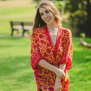 May include: A vibrant, knee-length kimono-style robe with a floral pattern in red and yellow. The robe has a contrasting red trim and is worn outdoors on a sunny day. The garment is lightweight and appears to be made of a flowing fabric.